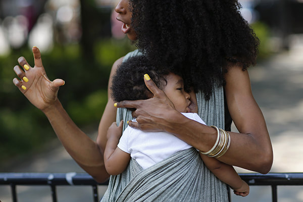 Babywearing can help with breastfeeding - even if you're not breastfeeding while babywearing. For more reasons why you should do babywearing, visit adjoyn.com/news [Image is a photo of a woman with brown skin wearing a baby on her front who is also breastfeeding. The woman is gesturing to someone off camera.]