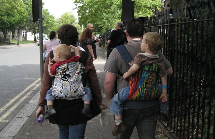 Baby carriers go anywhere! For more reasons why you should use a baby carrier, visit adjoyn.com/news [Image of two adults faced away from the camera showing the toddlers carried on their backs in buckle carriers. The adults are walking down a busy sidewalk.]