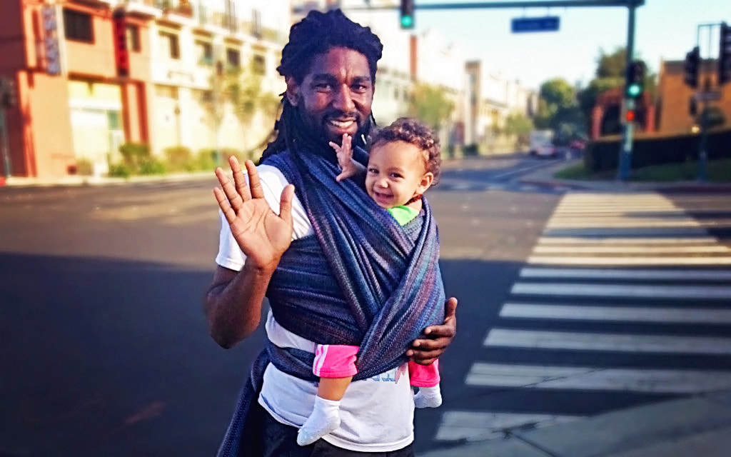 That baby is getting SO SMART. Babywearing helps make babies smarter, and it has a whole bunch of benefits for the wearer, too. More at adjoyn.com/news [Image of a man with dark skin wearing a toddler on his front in a woven wrap. He's standing near a street and waving; both the adult and toddler are smiling at the camera.]