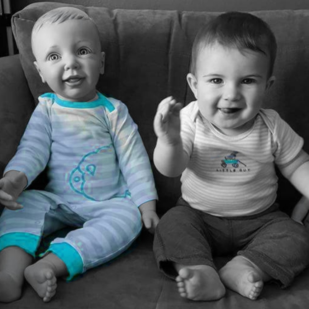 Meet the Fabies! Fake babies or babywearing demo dolls [Image of two babies sitting on a couch. The baby on the left is made of plastic with white skin and wearing a blue one piece jumper. The baby on the right is a real human 9 month old with white skin who is smiling at the camera.]